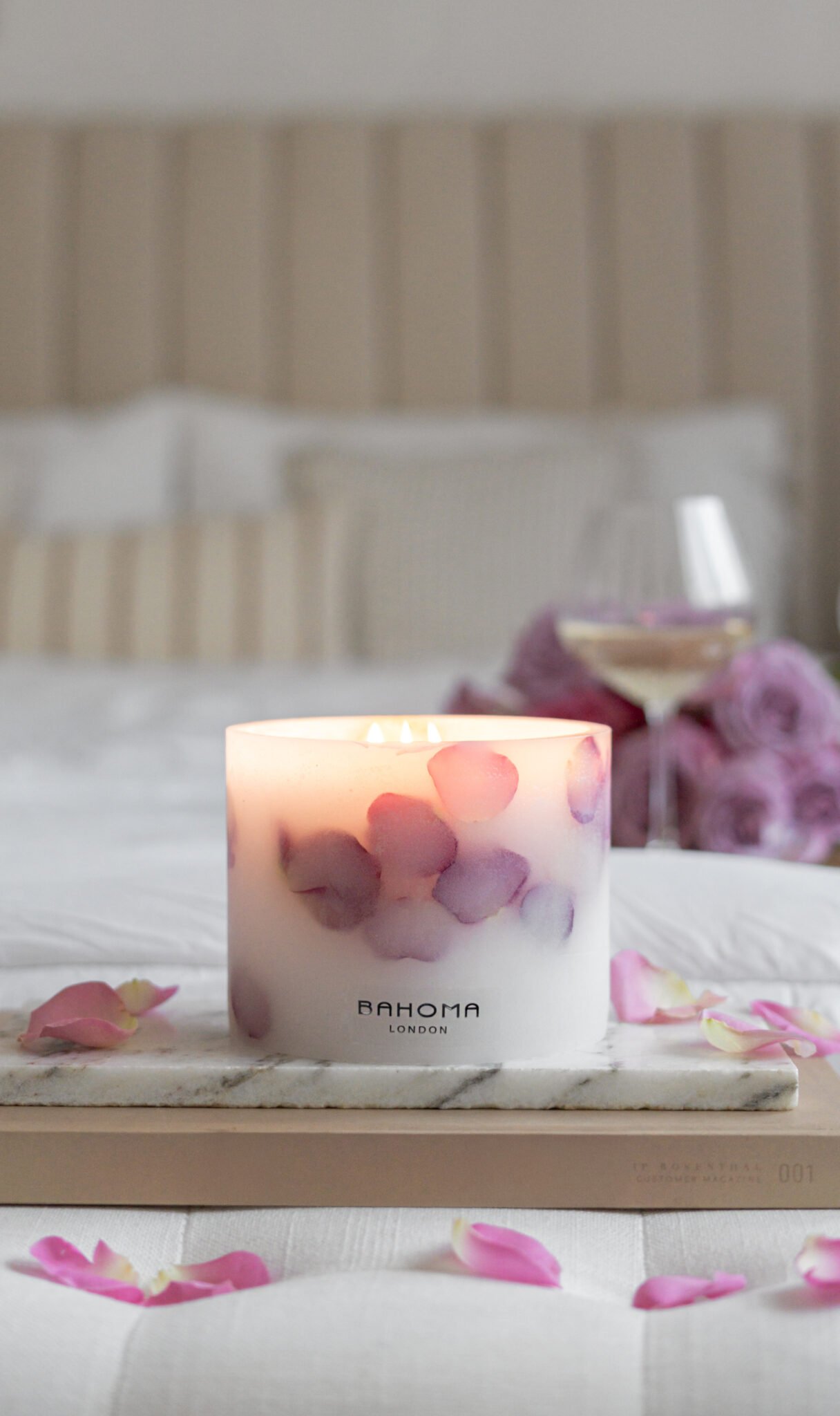 Grande Botanical Candle made with real rose petals on a side board with a bed and glass of wine in background