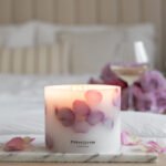 Grande Botanical Candle made with real rose petals on a side board with a bed and glass of wine in background