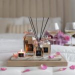 Velvet Rose - 500ml Fragranced Octagonal Diffuser with other size diffusers and travel candle arranged on a tray on a bed with rose petals scattered around.