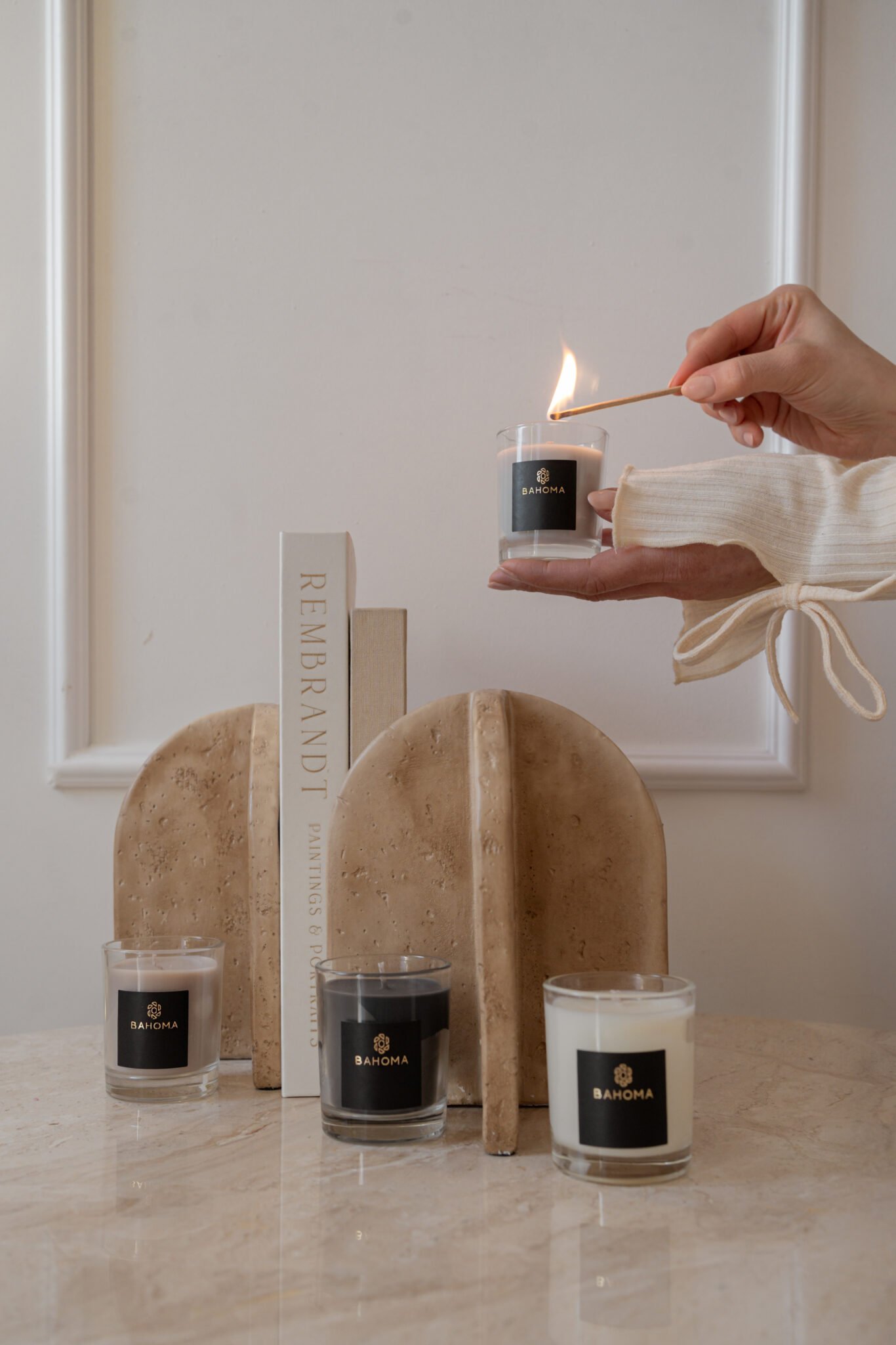Hand lighting a Bahoma scented travel candle from a 3-piece gift set; luxury home fragrance display with neutral decor and books.