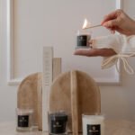 Hand lighting a Bahoma scented travel candle from a 3-piece gift set; luxury home fragrance display with neutral decor and books.